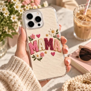 May include: A clear phone case featuring a cream-colored fabric insert embroidered with the word MAMA in pink, white, and burgundy block letters, accented with small embroidered daisies, green leaves, pink flowers, hearts, and gold stars.