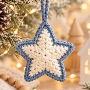 May include: A star-shaped crochet ornament featuring a cream-colored center with a textured stitch pattern, bordered by a thick blue yarn edge. The ornament includes a matching blue crocheted loop at the top for hanging, creating a two-tone design.