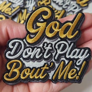 May include: Embroidered patch with the phrase "God Don't Play Bout' Me!" in gold and white thread on a black background. The patch is held in a hand, with other similar patches visible.