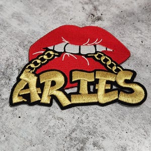 May include: A red and gold embroidered patch with a red lip design and a gold chain around it. The word "ARIES" is written in gold below the lip.