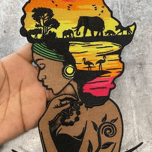 May include: Embroidered patch featuring a woman with an African continent silhouette as her hair. The continent is filled with a sunset scene with elephants, birds, and flamingos. The woman has a green head wrap and a floral tattoo on her arm.