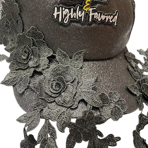 May include: A black trucker cap with a glittery finish, decorated with black lace floral appliques. The hat displays the text "BLESSED & Highly Favored" in white and yellow lettering.