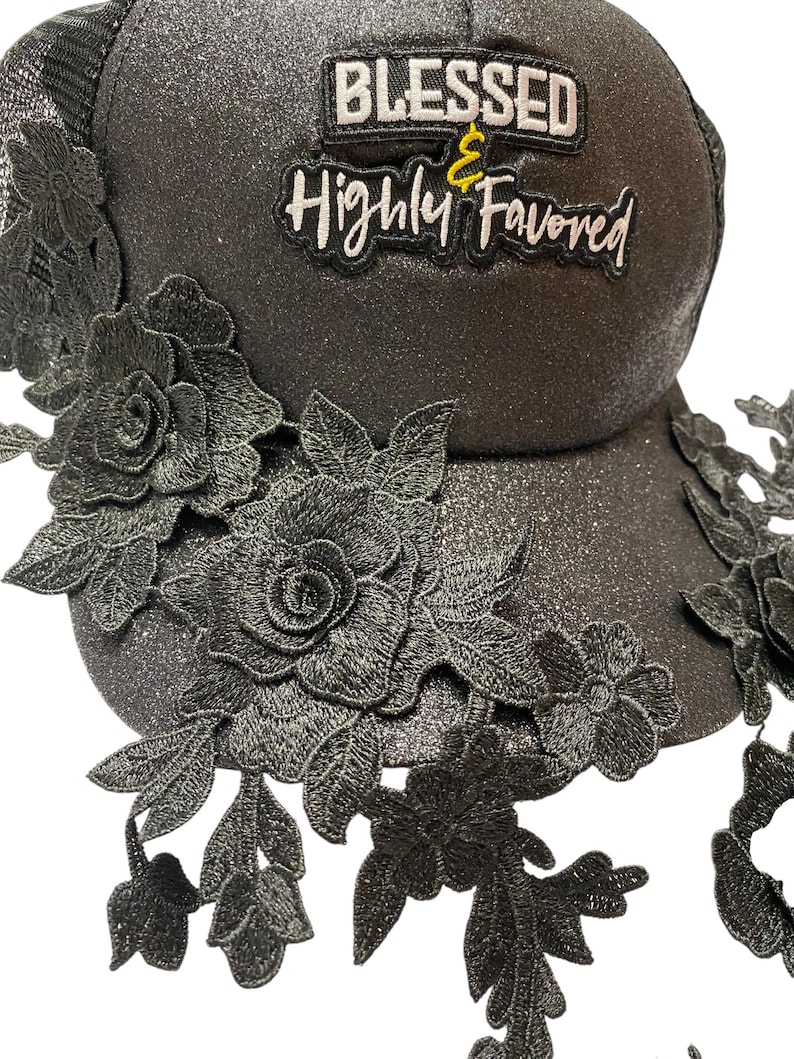 May include: A black trucker cap with a glittery finish, decorated with black lace floral appliques. The hat displays the text "BLESSED & Highly Favored" in white and yellow lettering.