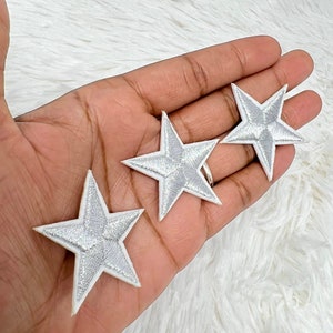 May include: Three silver star-shaped embroidered patches with white borders. The patches are arranged on a white, textured surface. The stars have a metallic sheen and are likely for decorative purposes.