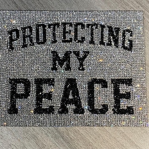 May include: A rectangular, silver-toned canvas covered in sparkling rhinestones. Bold, black lettering spells out "PROTECTING MY PEACE" across the surface, creating a glamorous and motivational piece of art.