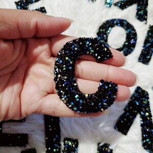 Peut inclure: Gros plan d'une lettre C noire en strass iris&eacute;s. La lettre est tenue dans une main sur un fond blanc flou.