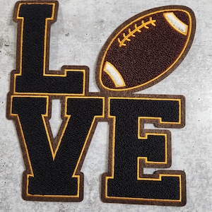 Puede incluir: Aplique de fieltro negro de la palabra "LOVE" con un balón de fútbol de fieltro marrón en el centro. Las letras están delineadas con hilo amarillo.