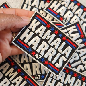 May include: A close-up of a stack of black fabric patches with white embroidered text that reads "Kamala Harris" and red and blue stripes. The patches have five white stars in a row with the number "47" below the stars.