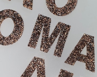 Può includere: Un set di cinque lettere dorate che compongono la parola "O-M-A-M" con una sola "O" al centro. Le lettere sono fatte di piccoli strass scintillanti.