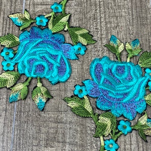 May include: Two turquoise blue embroidered rose appliques with green leaves and stems. The roses have a purple center and are surrounded by smaller turquoise blue flowers.