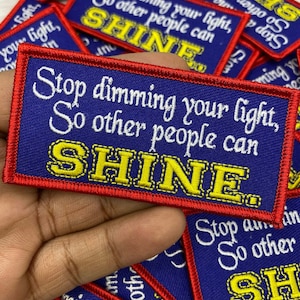 Op de afbeelding: Rechthoekige blauwe stoffen patches met rode randen en witte en gele geborduurde tekst. De tekst luidt: "Stop dimming your light, So other people can SHINE."
