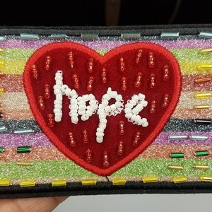 May include: A red felt heart with white beaded lettering spelling "Hope" on a multi-coloured striped background. The background is made of glitter and small, colourful beads.