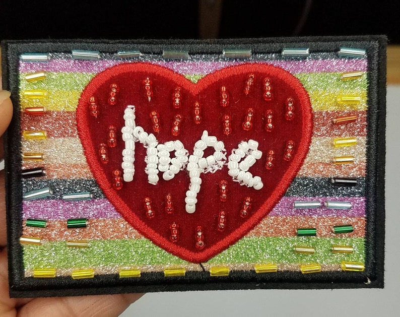May include: A red felt heart with white beaded lettering spelling "Hope" on a multi-coloured striped background. The background is made of glitter and small, colourful beads.