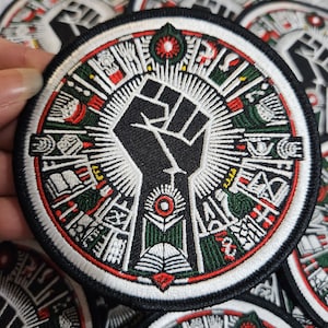 Può includere: Una toppa ricamata in bianco e nero con un pugno alzato al centro. Il pugno è circondato da un cerchio di simboli verdi, rossi e bianchi. La toppa è circolare e ha un bordo bianco.