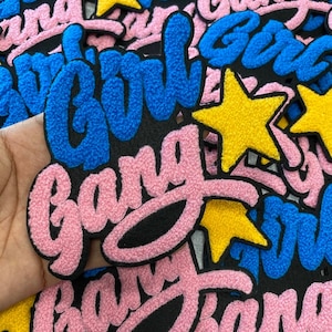 Op de afbeelding: Een close-up van verschillende roze, blauwe en gele viltpatches met de tekst "Girl Gang" in een gestileerd lettertype.