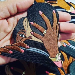 Puede incluir: Parche bordado con una mujer de cabello oscuro y piel morena, vestida con un atuendo negro y un reloj dorado. Su mano está levantada, mostrando un anillo de diamantes y esmalte de uñas rosa. El parche tiene forma de corazón.