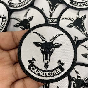 May include: A black and white embroidered iron-on patch with a goat head and the word "Capricorn" in a banner below. The patch is circular and has a black border.