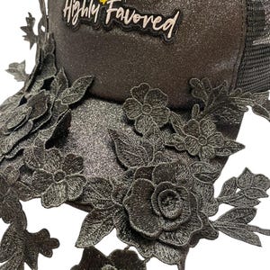 May include: Black trucker hat with a glittery finish, decorated with embroidered floral lace and the text "BLESSED & Highly Favored". The hat has a mesh back and a curved brim, a fashionable accessory.