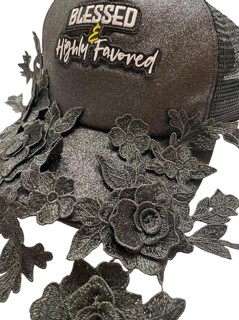 May include: Black trucker hat with a glittery finish, decorated with embroidered floral lace and the text "BLESSED & Highly Favored". The hat has a mesh back and a curved brim, a fashionable accessory.