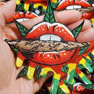 May include: A red embroidered patch with a pair of lips biting down on a brown marijuana joint. The lips are surrounded by green marijuana leaves and dripping with yellow and green paint.