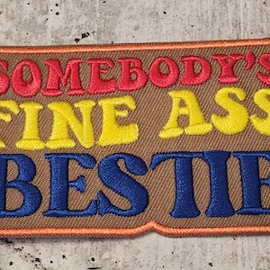 Puede incluir: Parche bordado con el texto "SOMEBODY'S FINE ASS BESTIE" en rojo, amarillo y azul. El parche tiene un fondo marrón con un borde naranja.