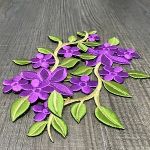May include: Purple and green embroidered flower applique with a vine design. The applique is made of fabric and has a detailed floral pattern.