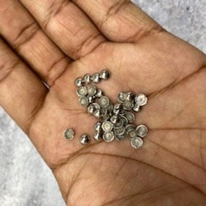 Könnte beinhalten: Ein Haufen silberfarbener Metallnieten, etwa 50 Stück, mit einer gewölbten Oberseite und einer flachen Unterseite. Die Nieten liegen auf einer Hand.