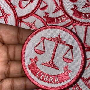 May include: A white and pink embroidered patch with a scale of justice and the word "LIBRA" in a banner.