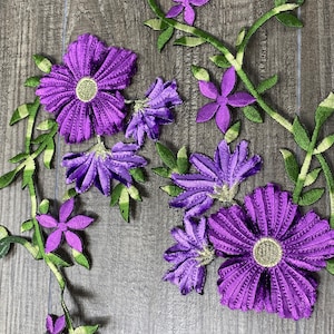 May include: Purple and green embroidered fabric flowers with a vine design. The flowers are in various sizes and shapes, with some having a more detailed design than others.
