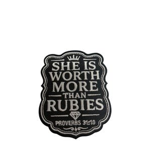 May include: A black embroidered patch with a scalloped border featuring white text that reads SHE IS WORTH MORE THAN RUBIES, a small crown icon above the text, a diamond icon below the word RUBIES, and the text PROVERBS 31:10 at the bottom.