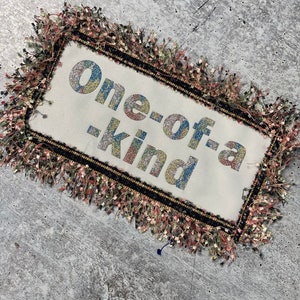 Puede incluir: Una almohada de tela blanca con un marco negro y ribete de flecos. La almohada tiene el texto "one-of-a-kind" en letras brillantes.