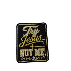 Può includere: Una toppa rettangolare ricamata nera con bordo dorato. Il testo "Try Jesus..." è scritto in corsivo dorato sopra una piccola croce, seguito da "NOT ME!" in lettere bianche, con un simbolo del giglio bianco centrato in basso.