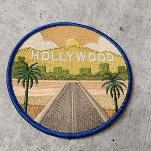 May include: Embroidered patch with a blue border, depicting a stylised landscape with palm trees, a road, and the text "HOLLYWOOD" in white.