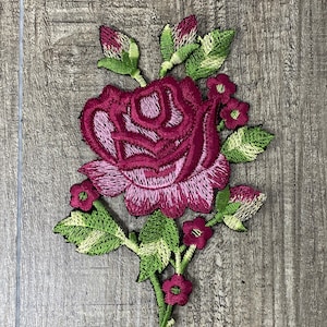May include: Embroidered floral patch featuring a large, open rose in shades of burgundy and pink, surrounded by green leaves and buds. The patch is likely for decorative purposes, such as embellishing clothing or accessories.
