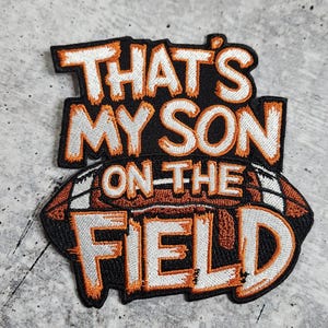 Może przedstawiać: Naszywka haftowana w kolorze pomarańczowym i białym z tekstem "That's My Son On The Field" i grafiką piłki nożnej.