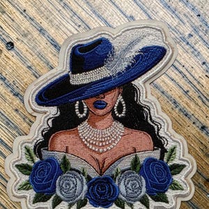 May include: Embroidered patch featuring a woman wearing a blue hat with a white feather, pearl necklace, and earrings. The design includes blue roses and a white dress. The patch has a white border and is on a wooden surface.
