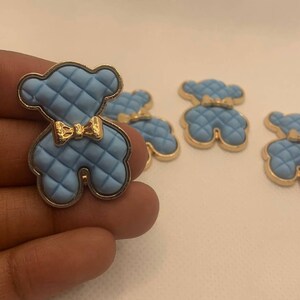 Op de afbeelding: Vijf blauwe en gouden knopen in de vorm van een teddybeer met een gestikte textuur en een gouden strik.