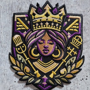 May include: A black and gold embroidered patch featuring a crowned woman with purple hair and gold earrings. The patch has a shield shape with a purple border and gold accents. The woman is surrounded by gold keys and leaves.