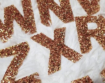 Può includere: Primo piano di un set di lettere di glitter dorate, tra cui le lettere M, N, X, R e Z. Le lettere sono disposte in uno schema casuale su una superficie bianca e soffice.
