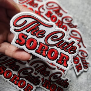 May include: Close-up of several white fabric patches with red embroidered text that reads "The Cute Soror".