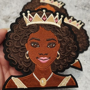 May include: Embroidered patch of a Black woman with brown curly hair wearing a gold crown with pink jewels, a gold necklace with a brown stone, and gold earrings.