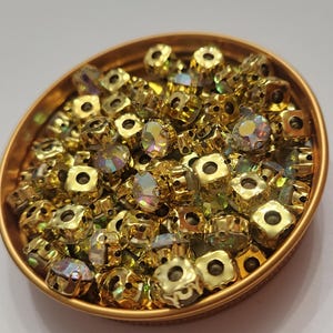 Peut inclure: Un ensemble de perles dorées de forme carrée avec des cristaux irisés à facettes. Les perles sont contenues dans une petite coupelle ronde dorée. Les cristaux reflètent un arc-en-ciel de couleurs.