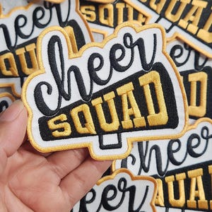 May include: Iron-on patch for a cheer squad, featuring a white background, black lettering, and a gold outline. The patch depicts a megaphone with the words "cheer squad" written on it.