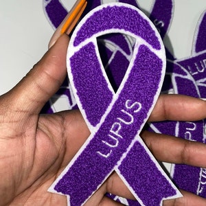 May include: Purple ribbon with white outline and the word "Lupus" embroidered in white. The ribbon is made of a fuzzy material.
