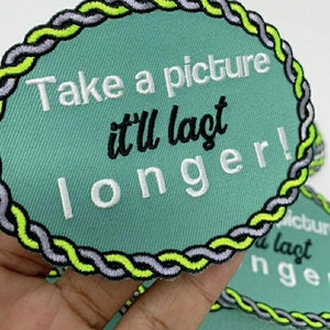Puede incluir: Un parche bordado verde con un borde de cuerda blanca y el texto "Take a picture it'll last longer!" en letras blancas y negras.