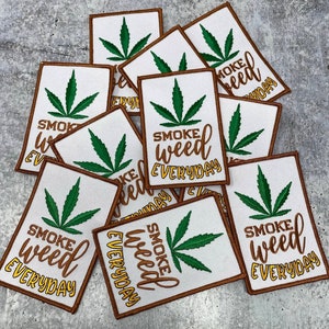 May include: A pile of white iron-on patches with a brown border. Each patch features a green marijuana leaf and the text "Smoke Weed Everyday" in yellow lettering.
