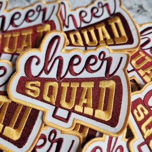 May include: A close-up of several white patches with red and gold embroidery. The patches feature the text "cheer squad" in a stylized font. The patches are perfect for adding a touch of school spirit to clothing, bags, or other accessories.