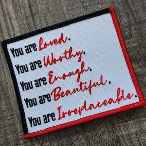 May include: A white patch with red stitching around the edges. The patch has black text that reads "You are Loved. You are Worthy. You are Enough. You are Beautiful. You are Irreplaceable."