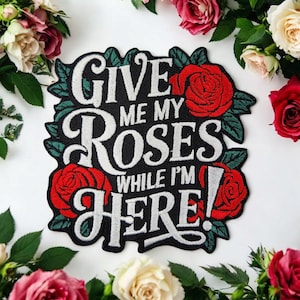 Può includere: Toppa nera con testo bianco che recita "GIVE ME MY ROSES WHILE I'M HERE!" circondata da rose rosse e foglie verdi. La toppa è circondata da un bordo di rose rosa, rosse e bianche.