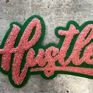 May include: A pink felt patch with the word "Hustle" embroidered in a cursive font on a green felt background.